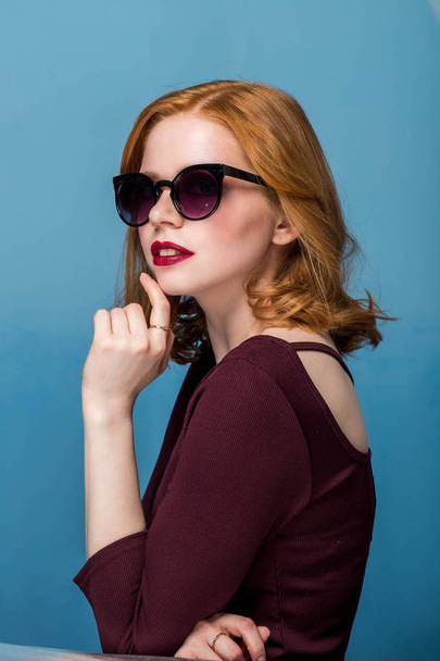 Redhead sexy beautiful girl with makeup and red lips in sunglasses on blue background. - Zdjęcie, obraz