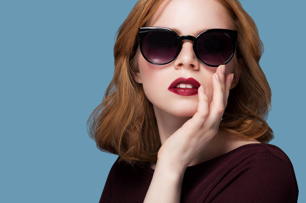 beautiful redhead girl in a maroon blouse red lipstick bright makeup in black sunglasses on a blue background closeup portrait - Photo, image