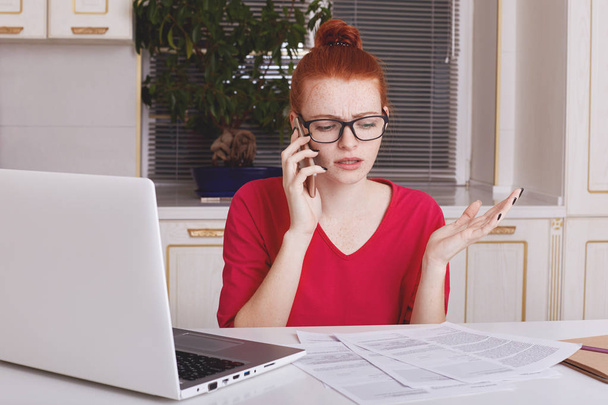 Puzzled ginger woman gestures actively as tries to explain something to employee via smart phone, sits at kitchen table, works with business documents and laptop computer. Occupation concept - Photo, Image