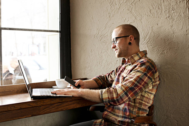 Hombre de negocios barbudo sonriente que usa ropa hipster casual usando computadora portátil y teléfono inteligente celular en la casa del café
. - Foto, Imagen