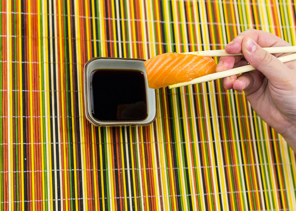 Arroz de sushi delicioso e uma fatia de mão de salmão segurando em paus de bambu é abaixado em uma tigela com molho de soja em um fundo multicolorido
 - Foto, Imagem
