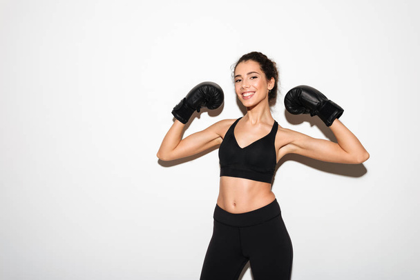 Smiling curly brunette fitness woman in boxing gloves showing biceps - Foto, imagen
