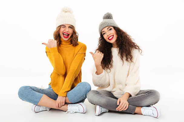Two playful girls in sweaters and hats sitting on floor - 写真・画像