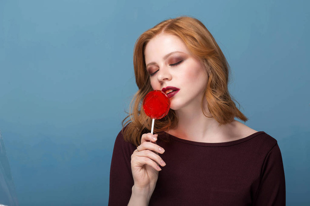 Stylish female model posing with lollipop and with closed eyes on blue background. - Фото, зображення