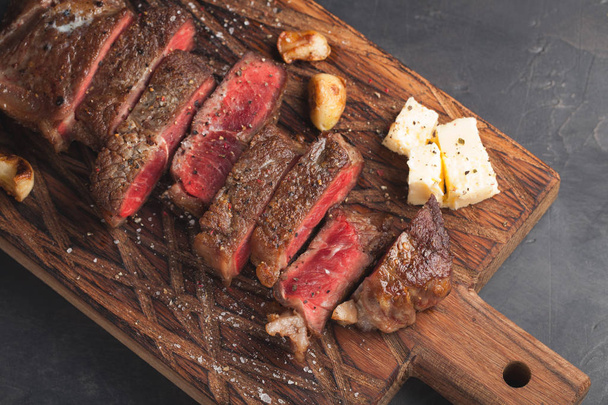 Nahaufnahme bereit, Steak New York Rinderrassen des schwarzen Angus mit Kräutern, Knoblauch und Butter auf einem Holzbrett zu essen. das fertige Gericht zum Abendessen auf dunklem Steingrund. Ansicht von oben - Foto, Bild
