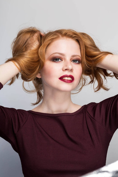 beautiful redhead girl with a smile, red lips, bright makeup looking at the camera happy hands and lifts the hair isolate portrait close-up - Photo, Image