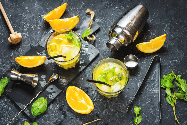 Холодный алкогольный летний цитрусовый коктейль с апельсином и мятой в стаканах и на темном каменном фоне. Инструменты для приготовления коктейлей, шейкер, стаканы, мятные листья
 - Фото, изображение
