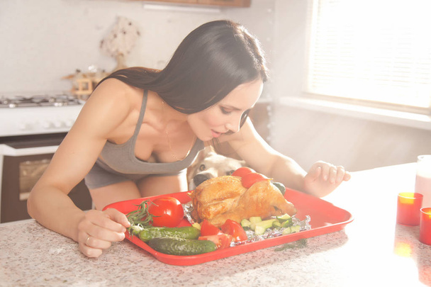 kaunis kunto brunette tyttö harmaa urheilu koti vaatteita valmistelee ja syö kanaa lähellä aamulla valo ikkuna hänen keittiössä kotona
 - Valokuva, kuva