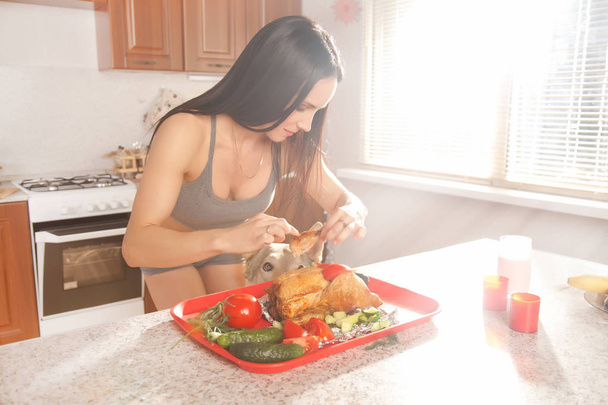 hermosa chica morena fitness en una ropa deportiva gris casa preparando y comiendo el pollo cerca de la ventana de luz de la mañana en su cocina en casa
 - Foto, imagen