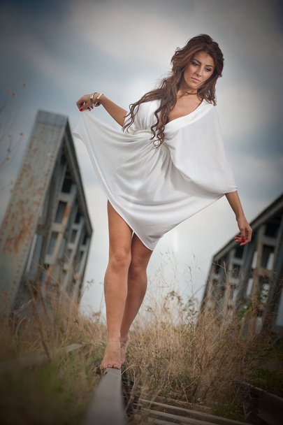 Attractive woman with short white dress and long hair standing on the rails with bridge in background. Fashion sexy girl with sexy body and long legs on the bridge posing in white dress  - Foto, Imagem