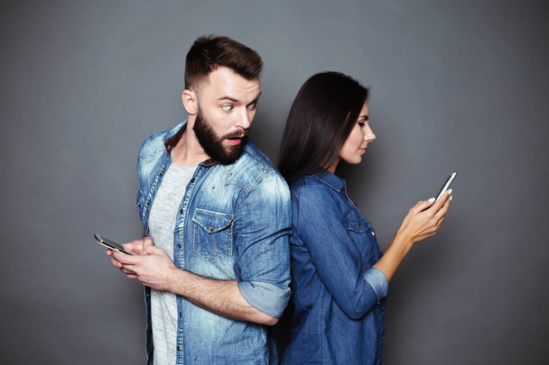 Beautiful young smiling couple in casual clothes standing back to back with phones in hands on a gray background isolated. - 写真・画像