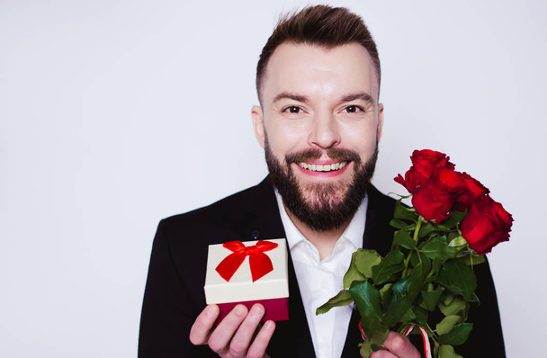 Handsome Smiling man in an elegant black suit with a gift box and red roses in hands looks at the camera on the white background isolated. - Zdjęcie, obraz