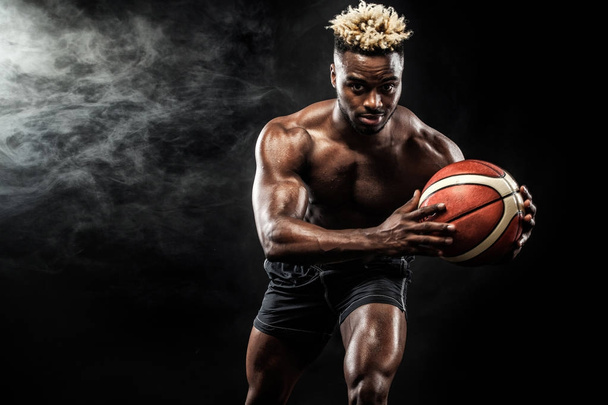 Portrait of afro-american sportsman, basketball player with a ball over black background. Fit young man in sportswear holding ball. - Fotoğraf, Görsel