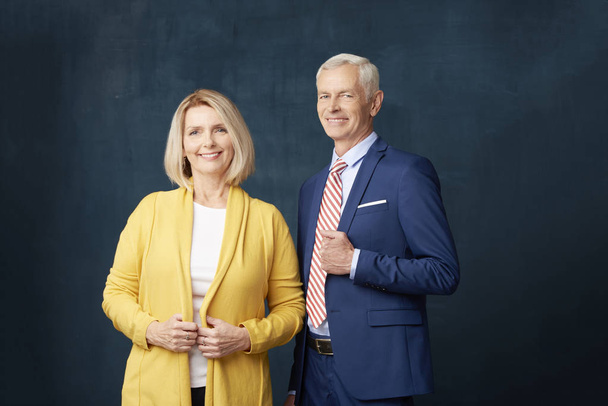 Studio shot of senior couple standing at dark background. Smiling mature woman looking at camera and elderly man wearing suit while standing next to her.  - Fotoğraf, Görsel
