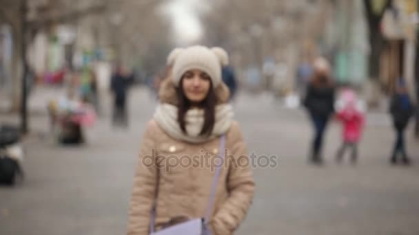 Happy girl in an anorak and a knitted hat calls by her mobile at a parking lot                               A splendid view of a cheery young woman in a knitted hat with balls, a fur anorak, calling  by her mobile near a white modern car in winter - Footage, Video