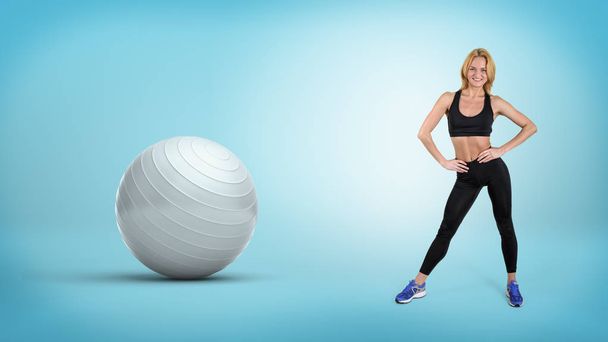 A young fit blonde woman stands with hands on her hips near a large silver exercise ball. - Foto, Imagem