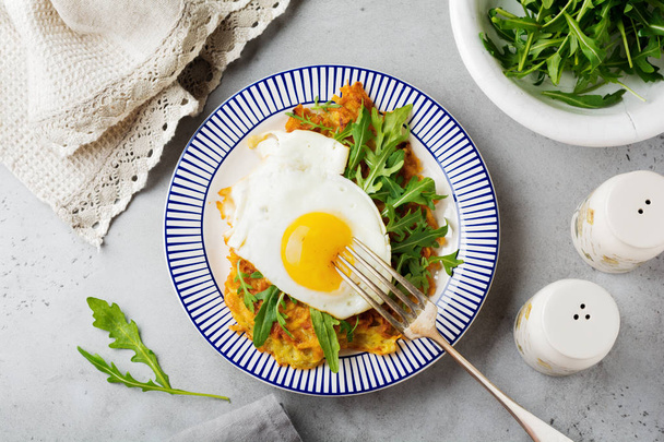 Fried egg with potato pancake, arugula and avocado on ceramic plate for breakfast on gray old concrete background. Selective focus. Top view. Copy space. - Photo, Image