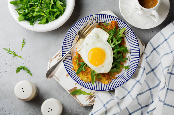 Fried egg with potato pancake, arugula and avocado on ceramic plate for breakfast on gray old concrete background. Selective focus. Top view. Copy space. - Fotoğraf, Görsel