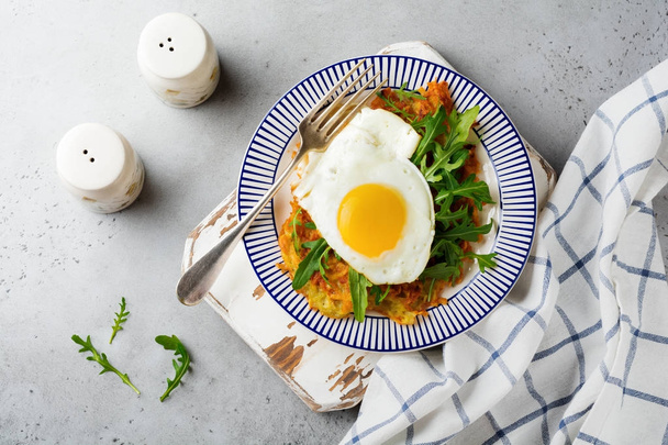 Fried egg with potato pancake, arugula and avocado on ceramic plate for breakfast on gray old concrete background. Selective focus. Top view. Copy space. - Valokuva, kuva