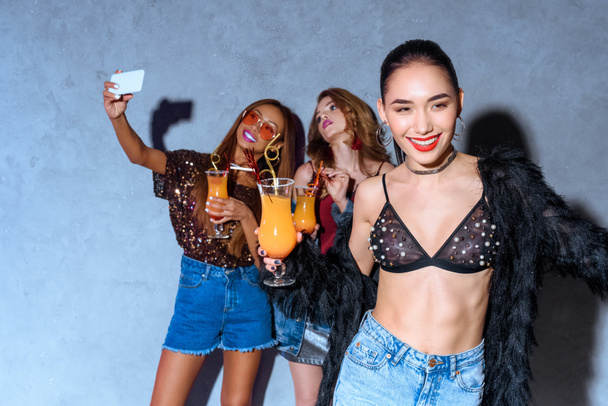sonriente elegante asiático mujer celebración de vidrio con cóctel mientras amigos beber cócteles y tomar selfie con teléfono inteligente detrás en la fiesta
  - Foto, imagen