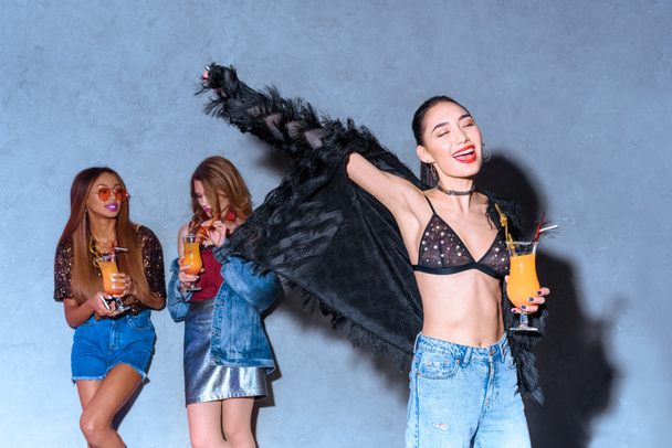alegre elegante asiático chica celebración de vidrio con cóctel mientras amigos bebiendo detrás en la fiesta
 - Foto, Imagen