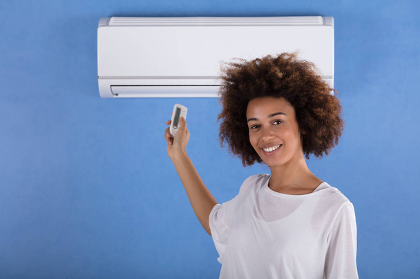 Young Woman Adjusting Air Conditioner Mounted On Blue Wall With Remote Control - Фото, изображение