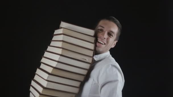 Человек в белой рубашке держит стопку книг в руках в темной комнате. Молодой бизнесмен с книгами в руках
. - Кадры, видео