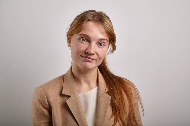 young redheaded woman with freckles and ponytail hairstyle wearing beige jacket and looking at camera with serious expression, student concept - Fotografie, Obrázek