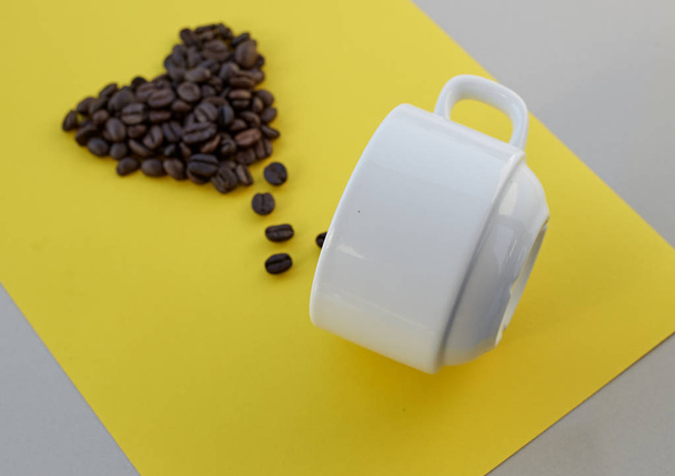 黄色の背景の上に白いカップとハートの形のコーヒー豆。バレンタイン コンセプト. - 写真・画像