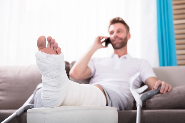 Happy Young Man With Broken Leg Sitting On Sofa Talking On Smartphone - Photo, Image