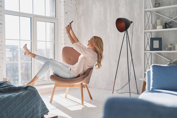 happy young woman taking selfie with smart phone while sitting in armchair at home - Fotoğraf, Görsel