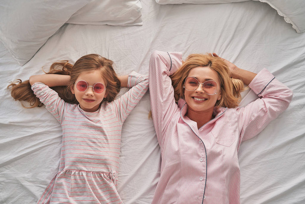 Top view of beautiful mother and cute daughter in sun glasses keeping hands behind head and smiling while lying on bed  - Fotoğraf, Görsel