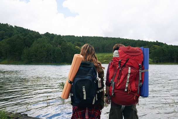 Rear view of couple with big backpacks standing at forest lake - Photo, Image