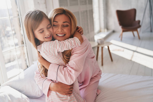 entzückendes kleines Mädchen, das seine Mutter umarmt, während sie zu Hause auf dem Bett sitzt - Foto, Bild