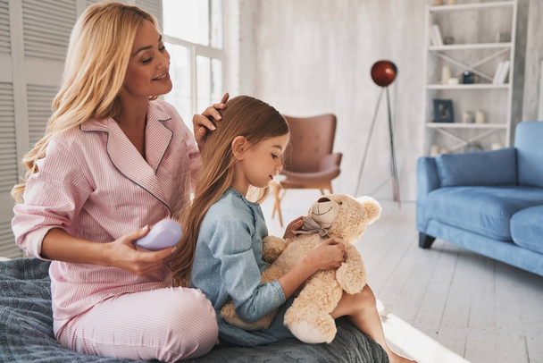 Beautiful young mother brushing her daughter hair while girl is holding teddy bear toy - Foto, Bild