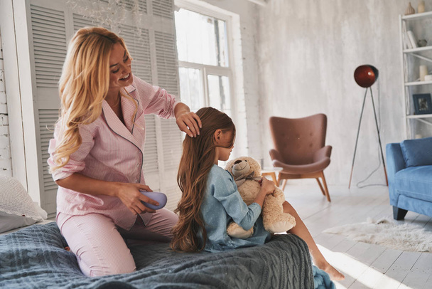 schöne blonde Mutter bürstet ihrer Tochter die Haare, während sie zu Hause auf dem Bett sitzt - Foto, Bild