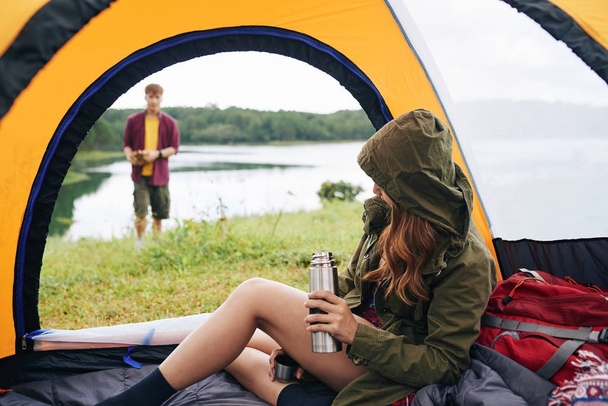 Young woman sitting in tent and drinking tea when her boyfriend is making campfire - Photo, Image