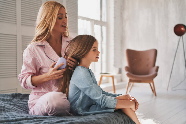 blonde mother brushing her daughter hair in bedroom - Photo, Image