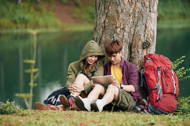 Vietnamese young travelers sitting under rives and reading something on tablet computer - Photo, Image