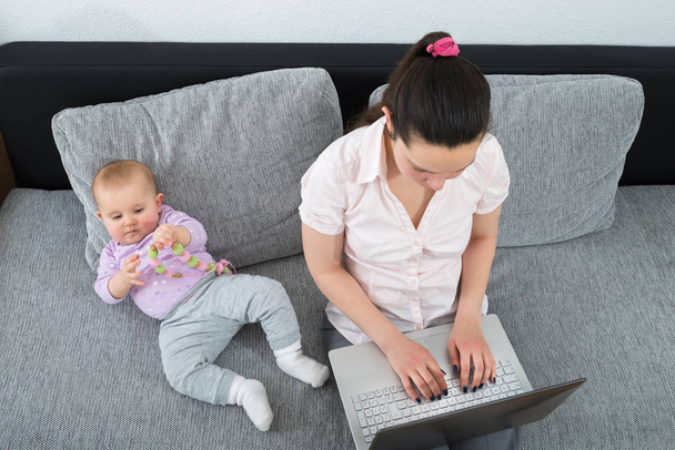 Woman Sitting On Sofa Using Laptop While Her Baby Playing With Toy - Photo, image