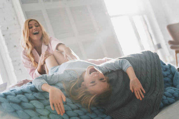 Beautiful blonde mother going crazy with her cute daughter in bed on knitted blanket - Photo, Image