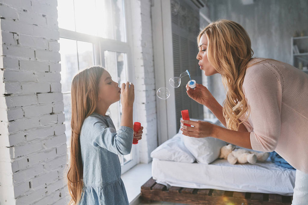 Madre e hija soplando burbujas de jabón mientras pasan tiempo en casa
 - Foto, Imagen