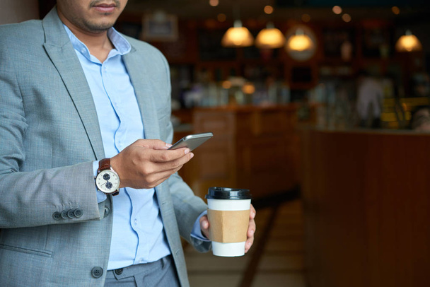Immagine ritagliata dell'uomo d'affari che beve caffè da asporto e controlla il telefono
 - Foto, immagini