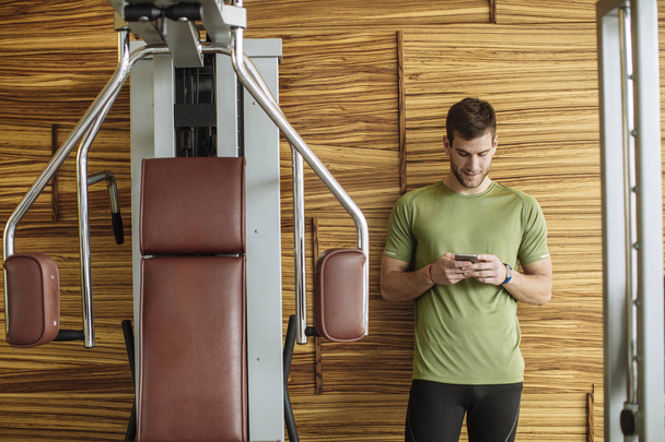 Handsome Caucasian sportsman typing on smartphone at gym. - Fotó, kép