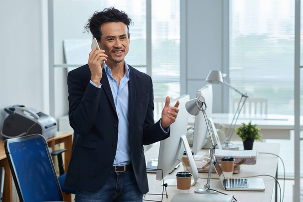 Sonriente hombre de negocios de carrera mixta hablando por teléfono en su oficina
 - Foto, imagen