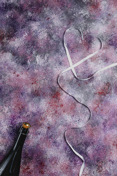 Draufsicht auf Champagnerflasche und herzförmiges Band auf lila Tischplatte, Valentinstag-Konzept - Foto, Bild