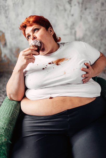 overgewicht vrouw zitten in de leunstoel en het eten van zoete cupcake, luiheid, obesitas en ongezonde voeding eten concept - Foto, afbeelding