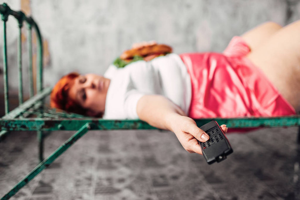 λίπος γυναίκα ξαπλωμένη στο κρεβάτι σκουριασμένο, τρώνε σάντουιτς και βλέποντας τηλεόραση, ανθυγιεινό τρόπο ζωής και παχυσαρκία έννοια - Φωτογραφία, εικόνα