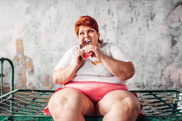υπέρβαρα γυναίκα κάθεται στο σκουριασμένο κρεβάτι και τρώει σοκολάτα, την τεμπελιά και την έννοια της παχυσαρκίας. Τρώγοντας ανθυγιεινά τρόφιμα - Φωτογραφία, εικόνα