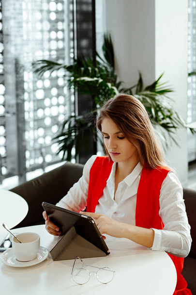 The strict business lady sits at a little table in cafe, uses the Internet on the tablet and has coffee. The woman in a white shirt and a red jacket during a lunch in business center. - 写真・画像